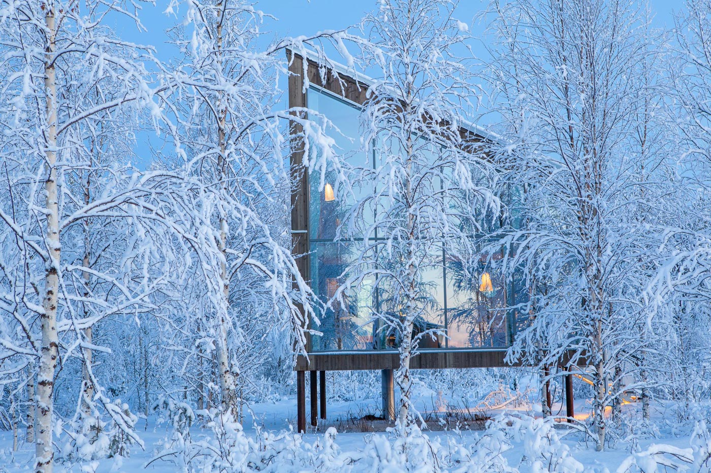 Eine Außenansicht eines Holzhauses und der schneebedeckten Birkenlandschaft. Secto Leuchten scheinen durch die großen Fenster.