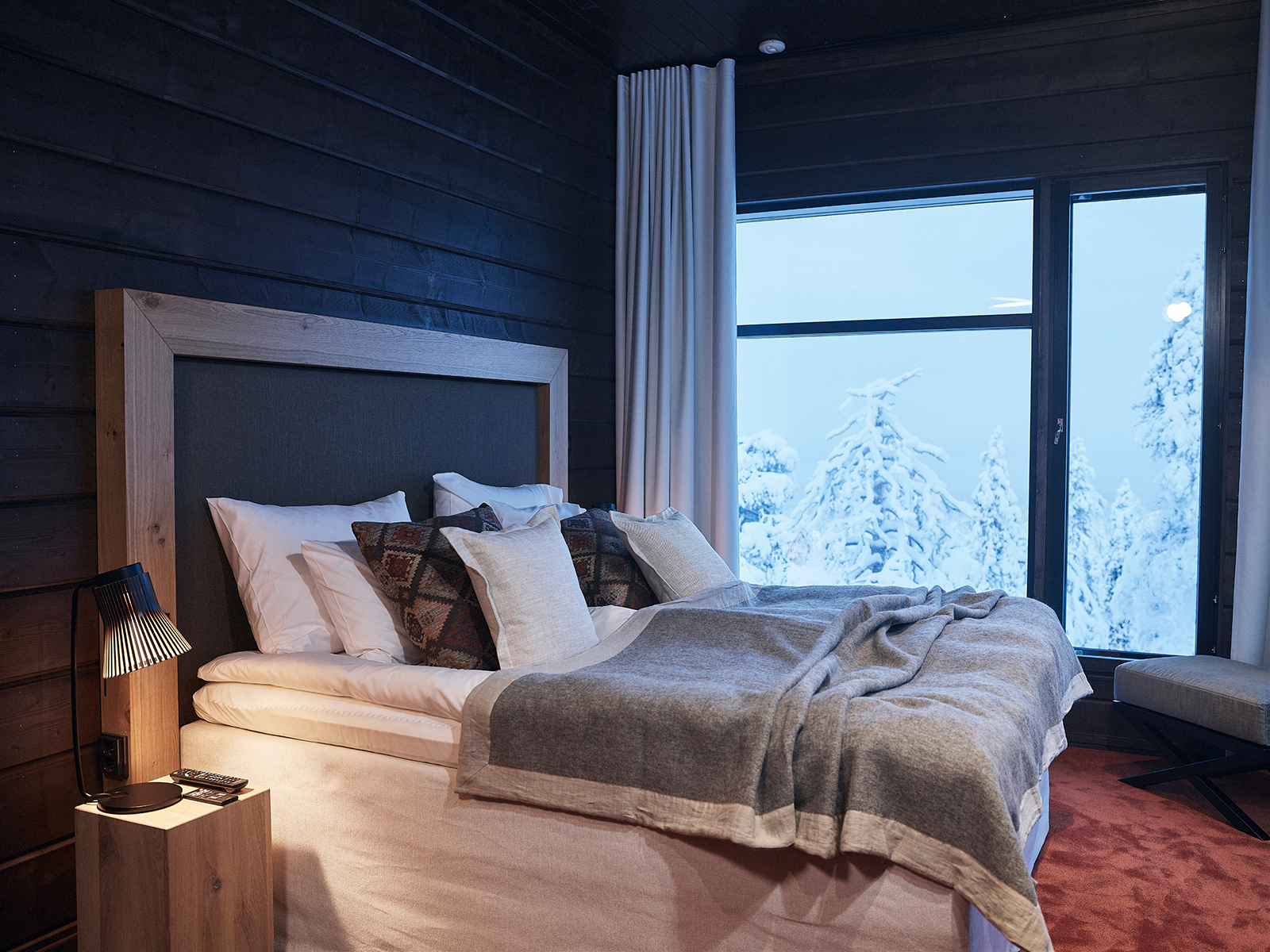 Ein Schlafzimmer mit einer Petite Tischleuchte auf dem Nachttisch, große Fenster zeigen schneebedeckte Bäume.