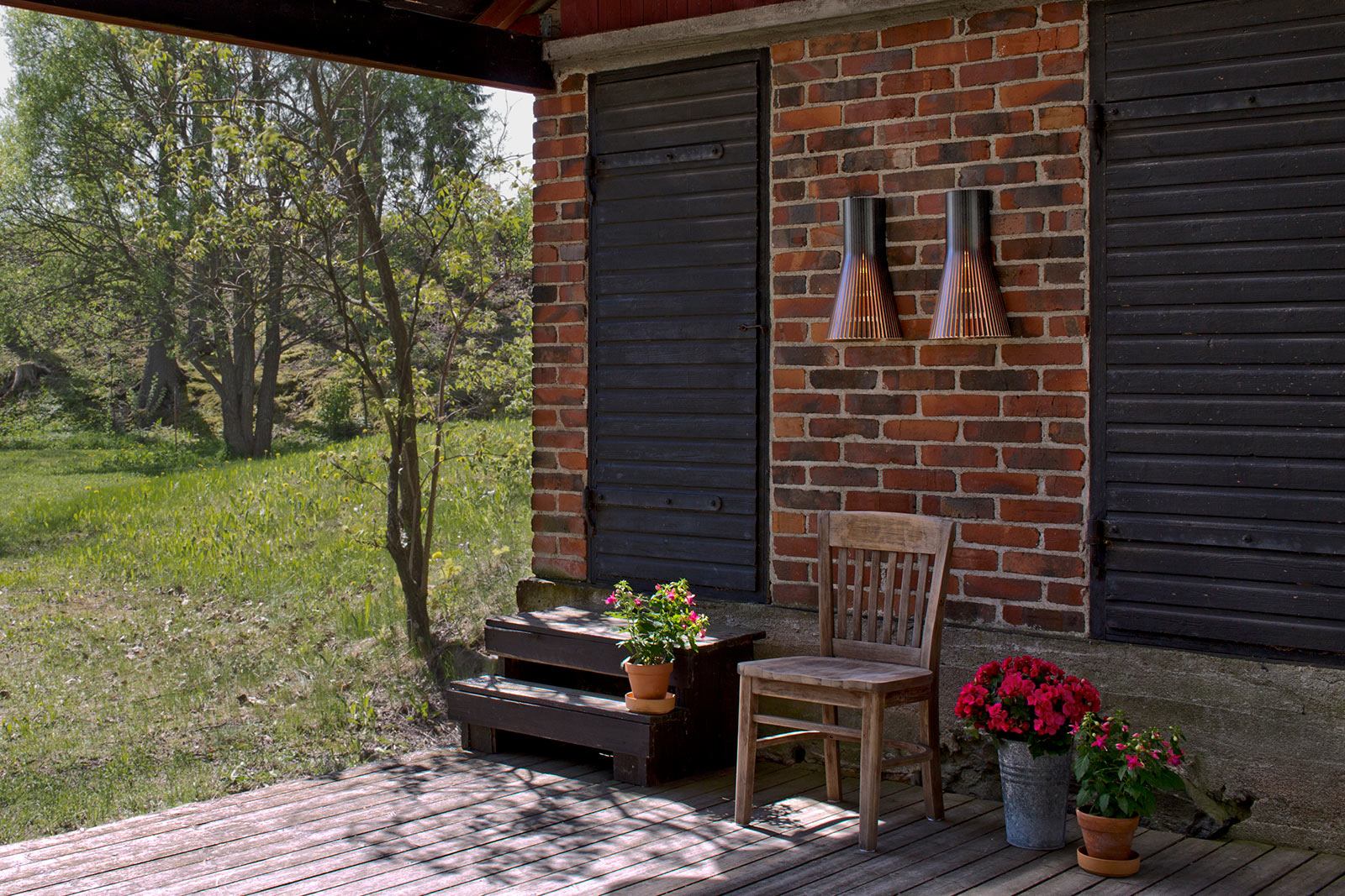 Eine überdachte Terrasse, Pflanzen und ein Stuhl vor einer Ziegelwand. Zwei schwarze Türen, dazwischen Secto Wandlampen.