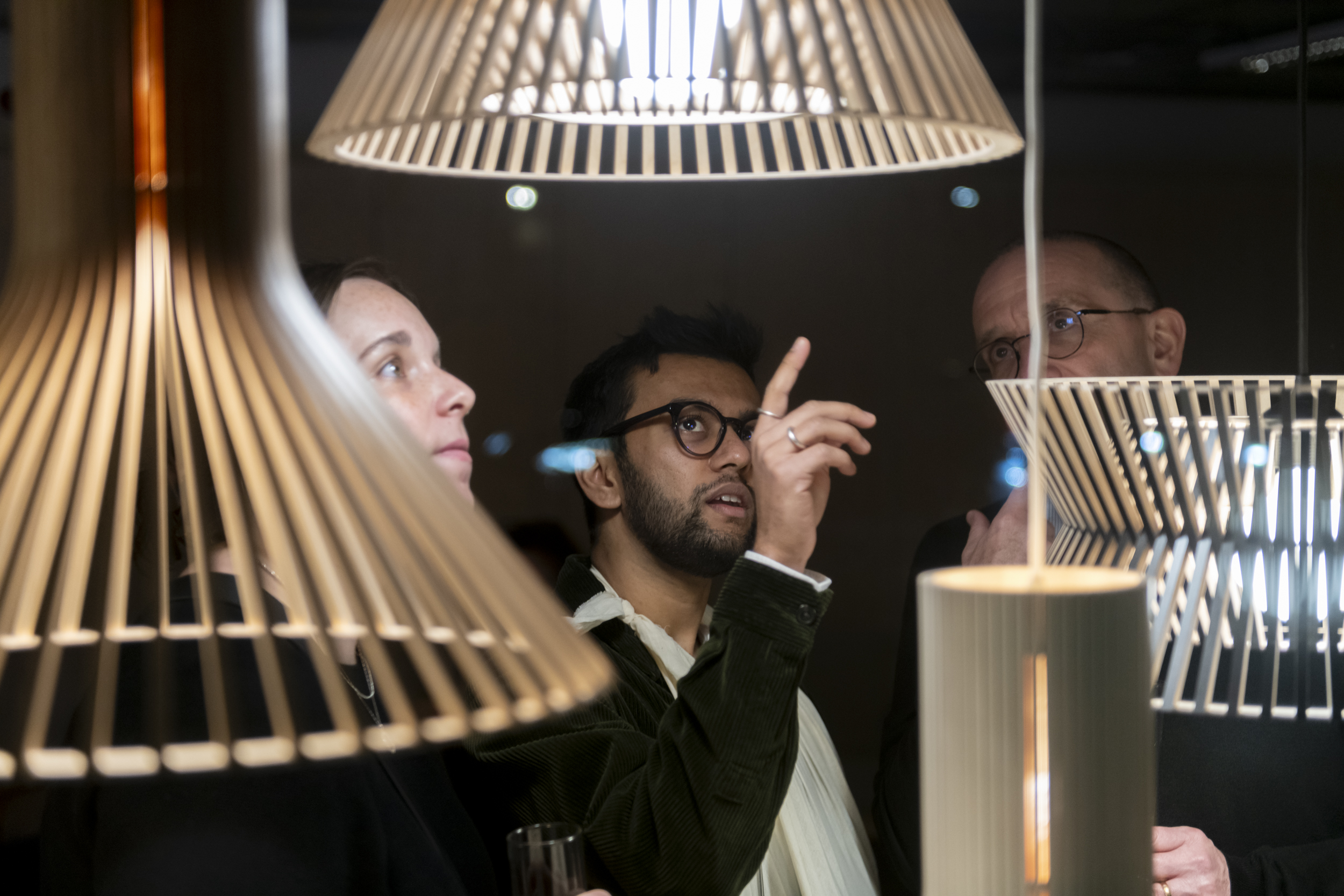 A woman and two men looking upwards at pendant lamps. The man in the middle points the uppermost lamp.