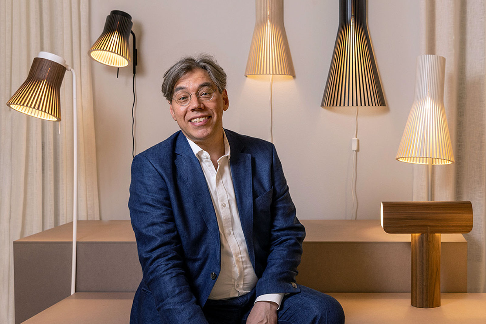 A man leans against a long conference table, lit by three pendant lights. White curtains in the background.