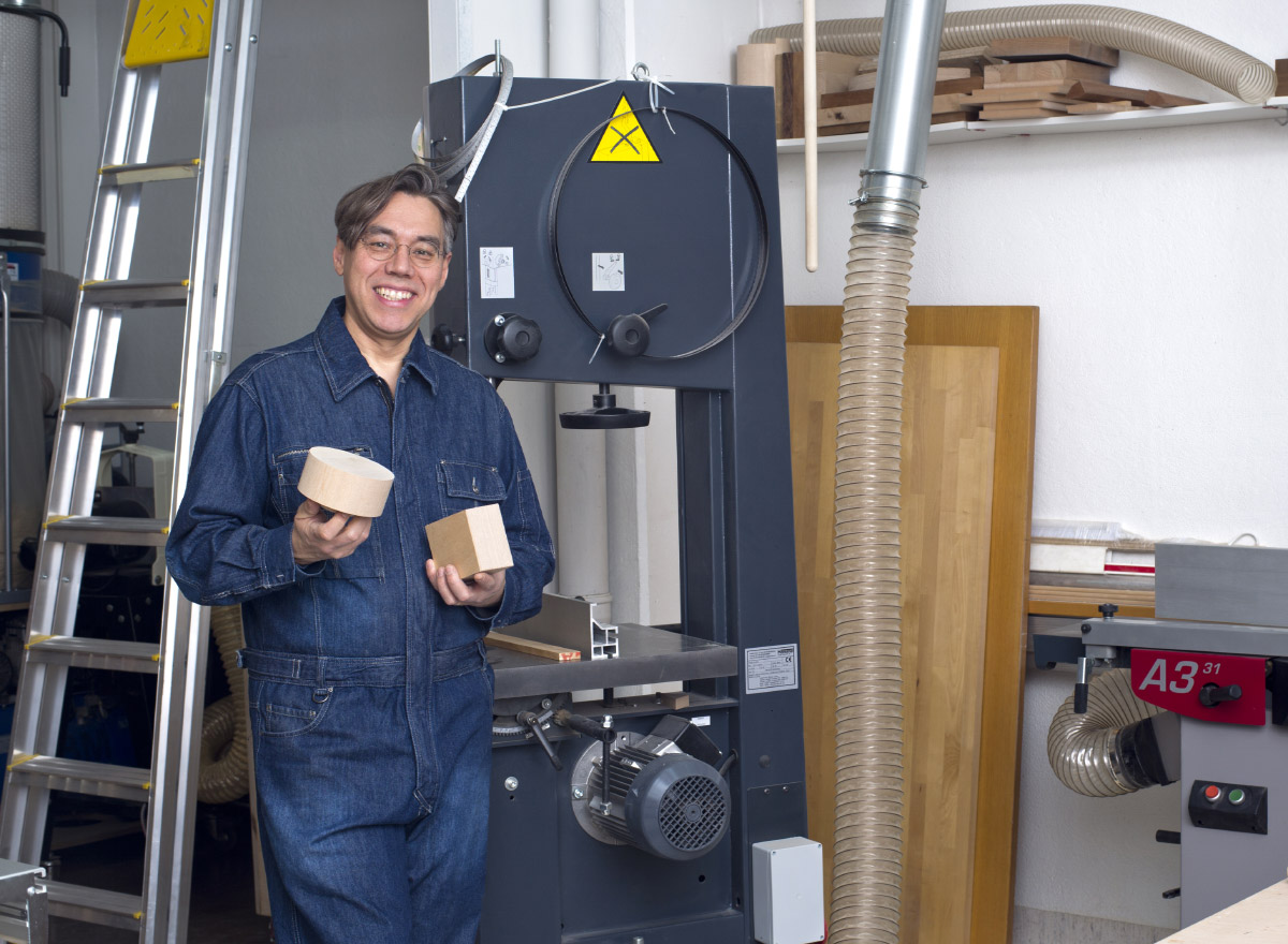The designer stands beside a woodworking machine, holding blocks of wood. 