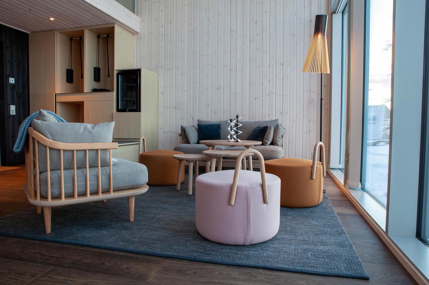 Ein Wohnzimmer mit hellen Möbeln, Holzwänden und großen Fenstern. Eine Secto Stehlampe steht rechts davon.
