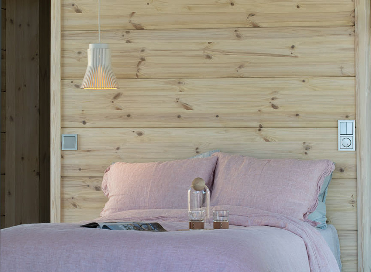 Ein Bett mit hellrosa Bettwäsche vor einer Holzwand. Eine Petite Pendelleuchte hängt daneben.