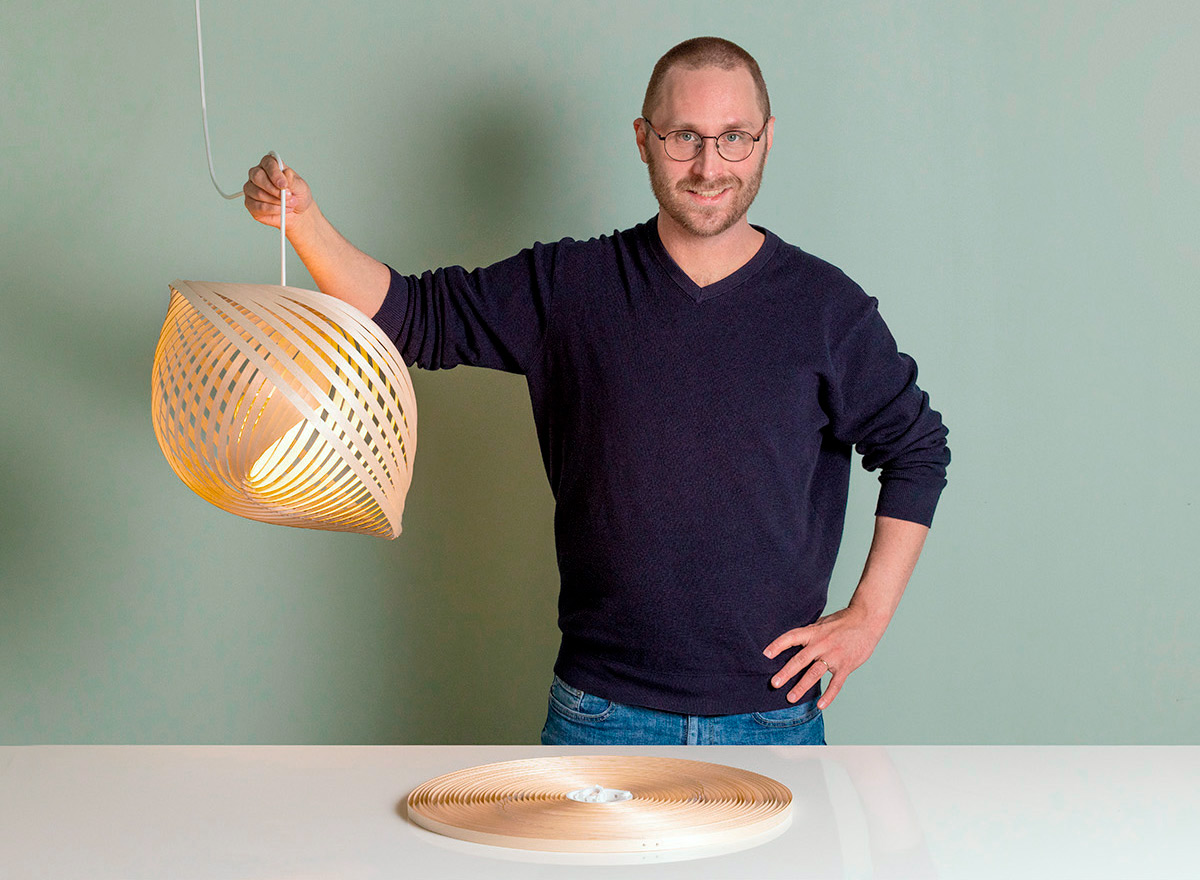 The designer stands behind the table, holding the cable of a pendant lamp. On the table, there is a disc made of plywood rings.