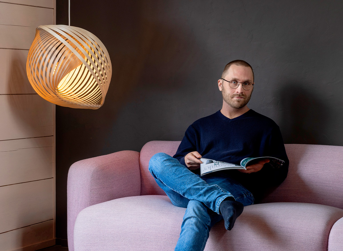 The designer sits on a sofa, holding a magazine. A lamp hangs in the corner.