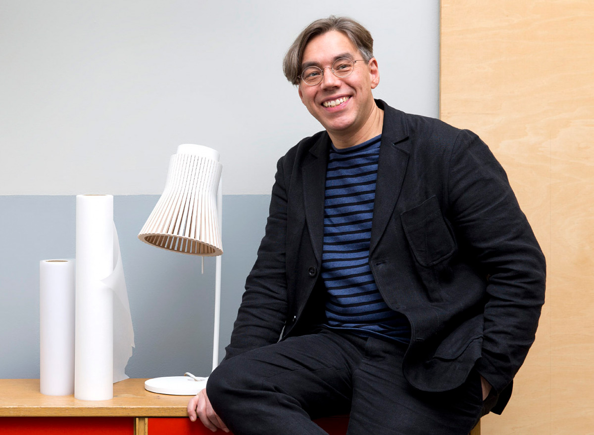 The designer leans on a table, with rolls of drafting paper and a desk lamp nearby.