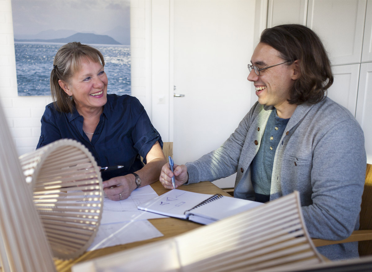 The owner and the designer are laughing at a table which is full of wooden design lamps and sketches.