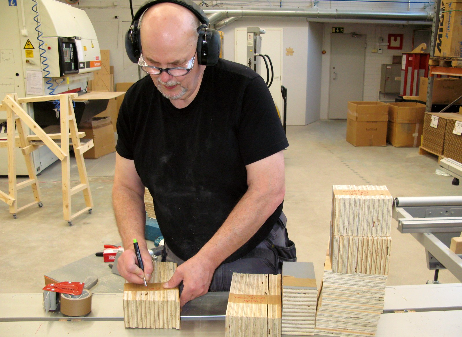 A man is drawing some marks on wooden blocks. He is wearing hearing protection and safety glasses.