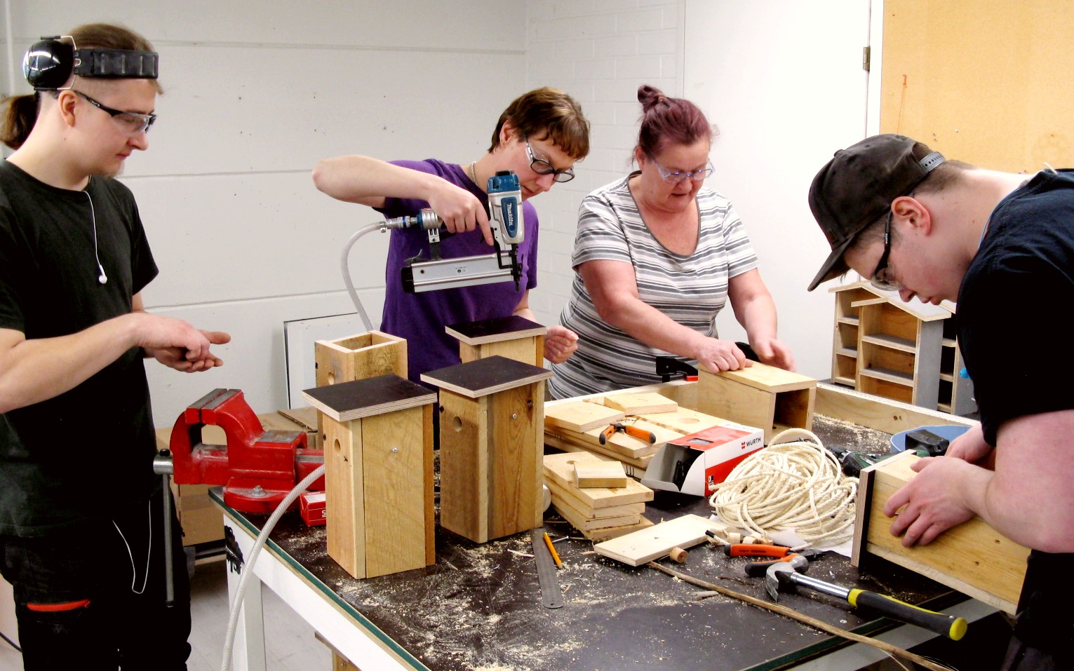 A desk with some self-made birdhouses in various stages. Four people are working on them.