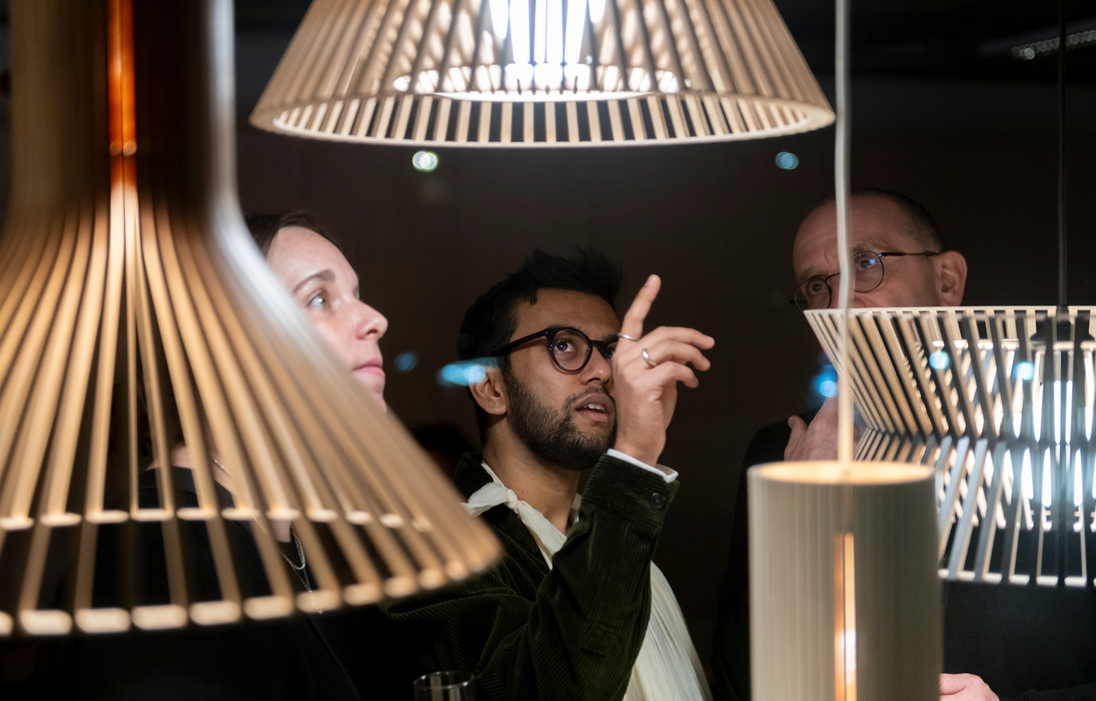 A woman and two men looking upwards at pendant lamps. The man in the middle points the uppermost lamp.