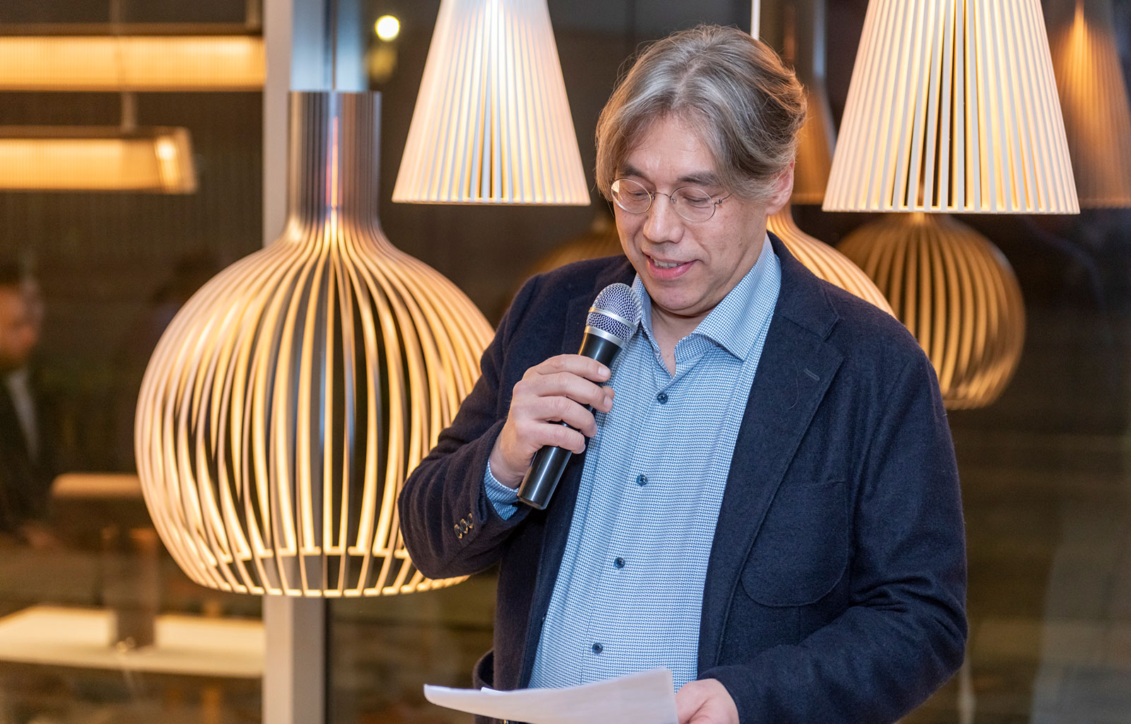 Un homme souriant avec un microphone et des notes dans les mains devant des lampes en bois.