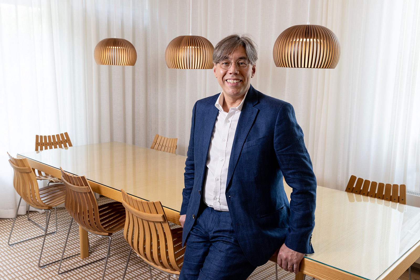 A man leans against a long conference table, lit by three pendant lights. White curtains in the background.