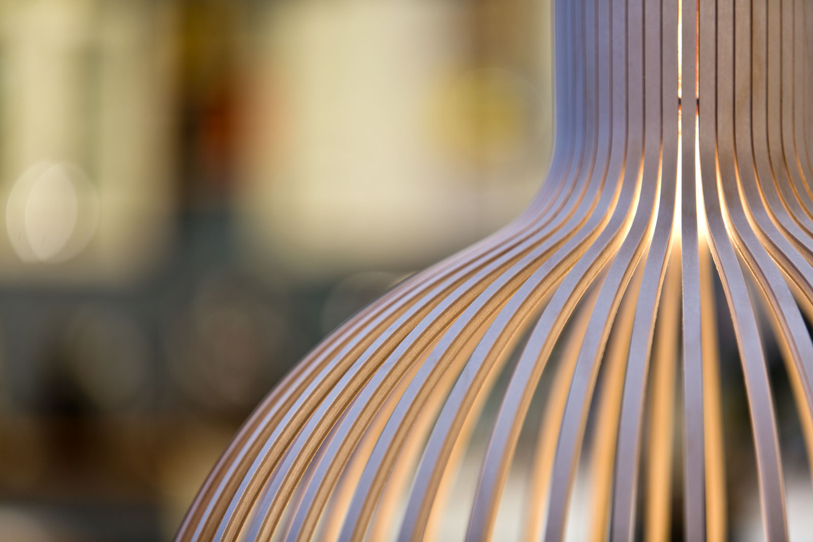 Close-up of a wooden pendant lamp with dense upper slats hiding the light source and lower slats diffusing the light.