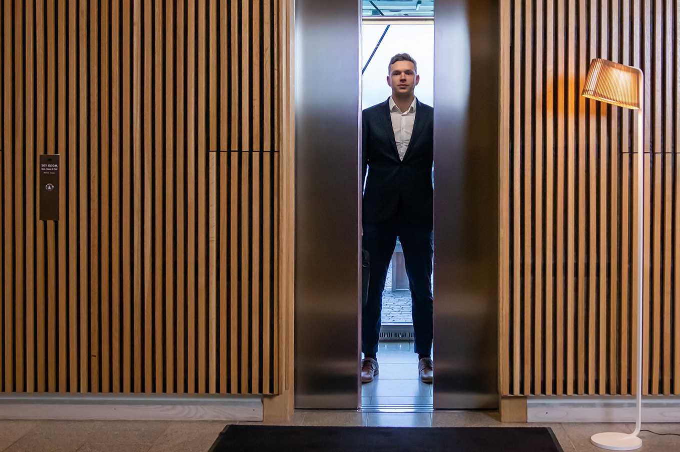 A man standing in an elevator with open doors. The Owalo floor lamp is placed against a wooden wall.