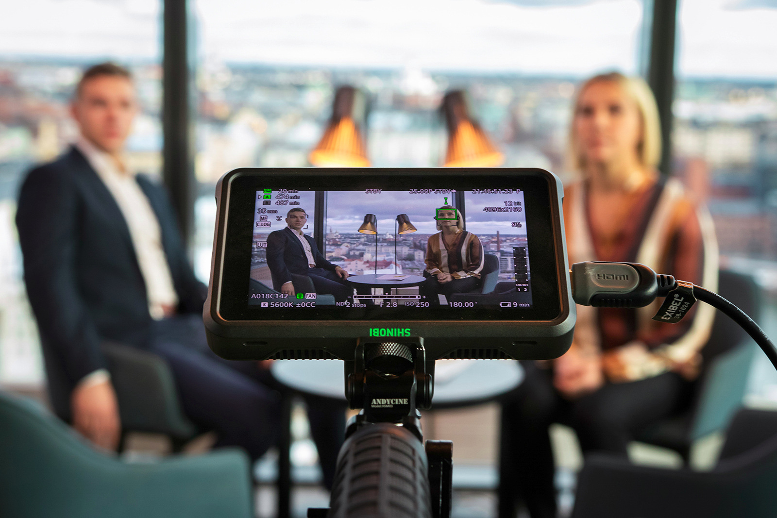 A monitor showing the background: a man and a woman sitting at a small table. Floor lamps and Helsinki in the background.