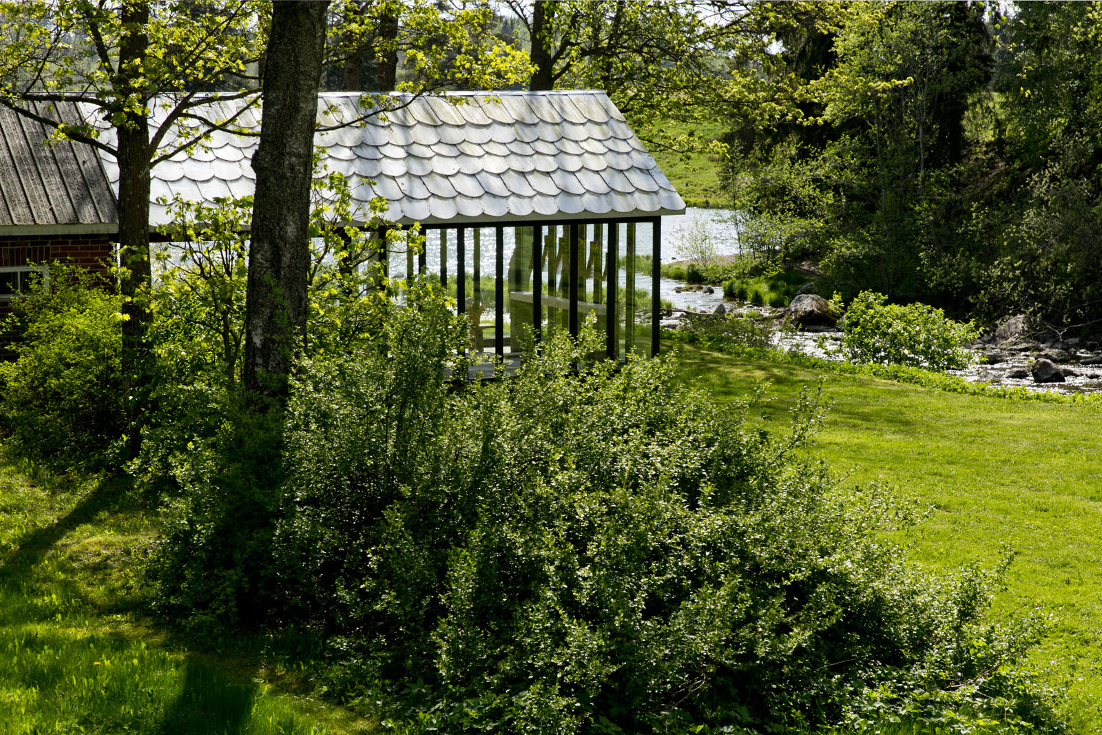 A brick house with a glass porch on the left, a small river flows into a lake from the right through the garden.