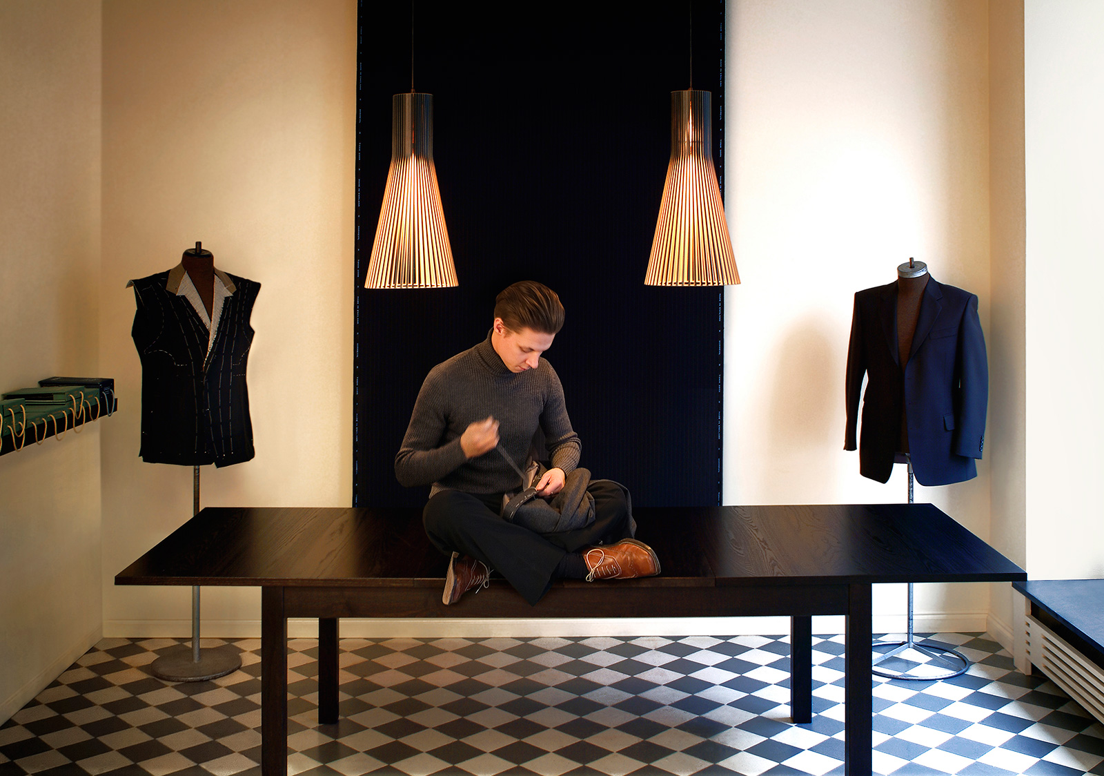 A taylor sitting on a table in a sewing room, working on a jacket. Two black Secto pendant lamps light up the room.