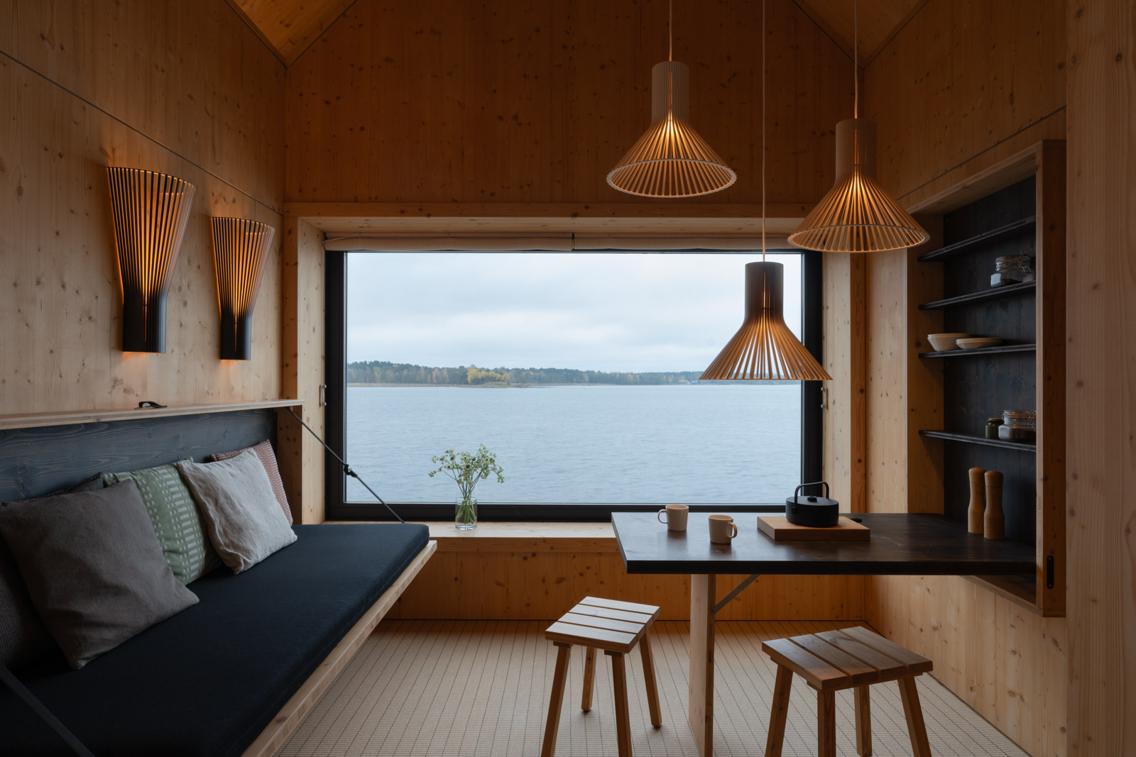 Wooden Majamaja cabin with Puncto pendant lamps and Secto wall lamps, natural light from the sea-facing window creating a calm Nordic atmosphere.