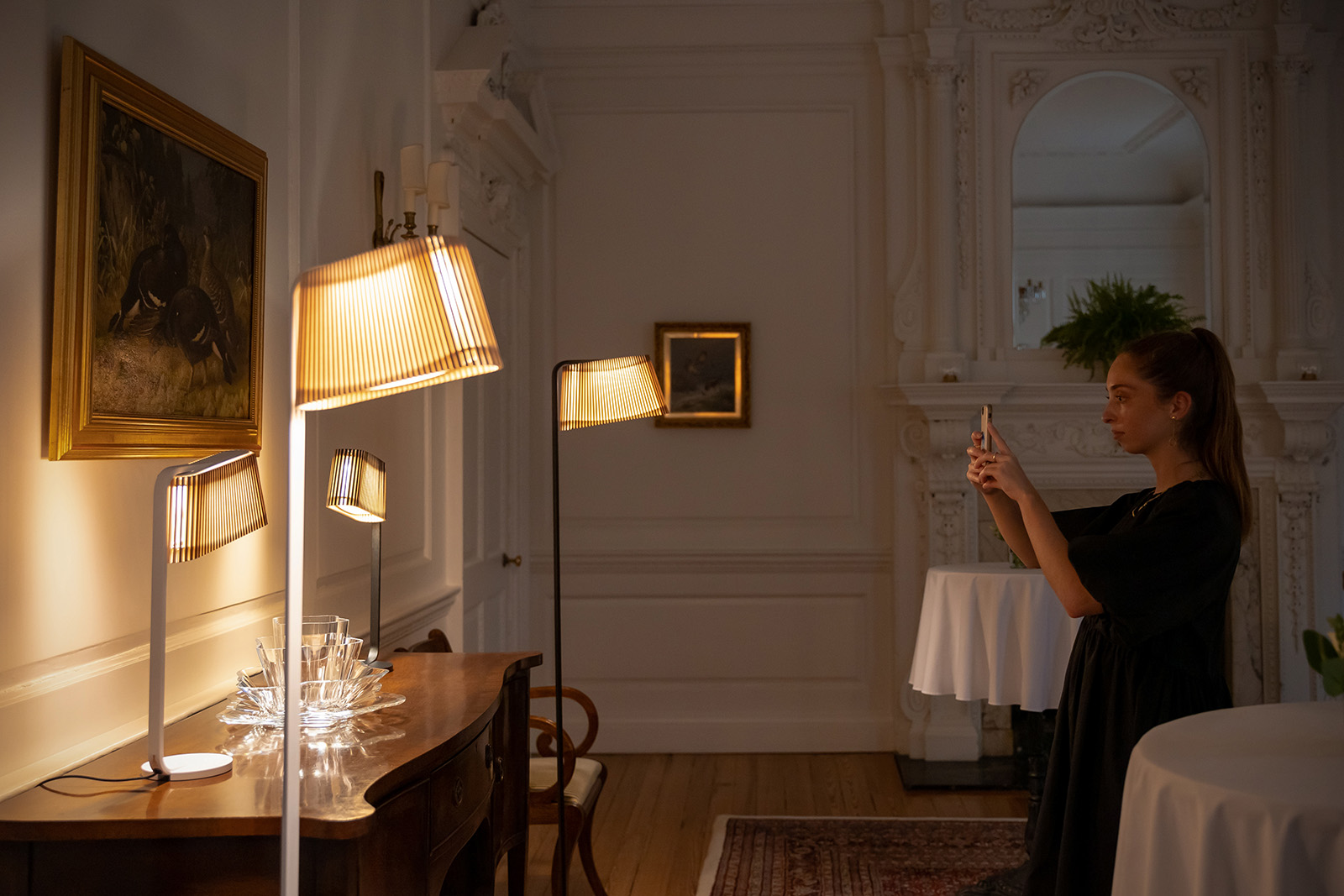 A woman taking a photo of a painting, a sideboard and floor and table lamps.