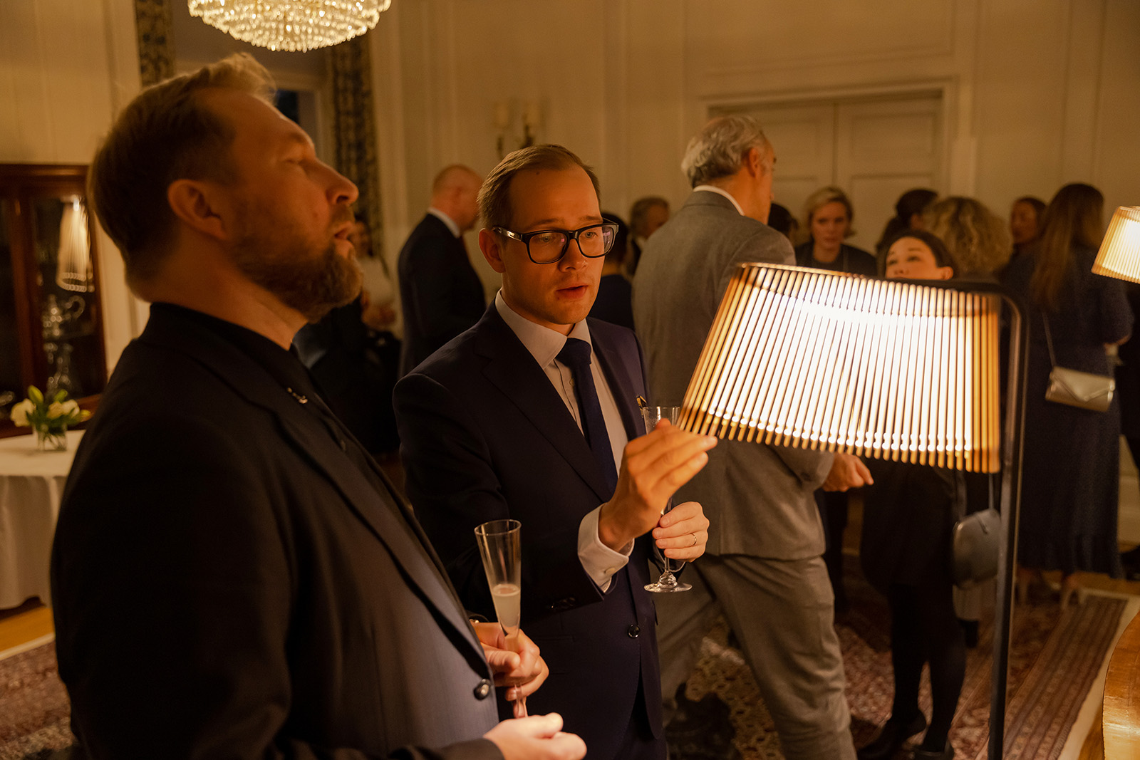 Two men examining a wooden lamp shade. More people in the background.